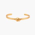 Gold cuff bracelet with twisted rope texture and decorative knot centerpiece on cream background.