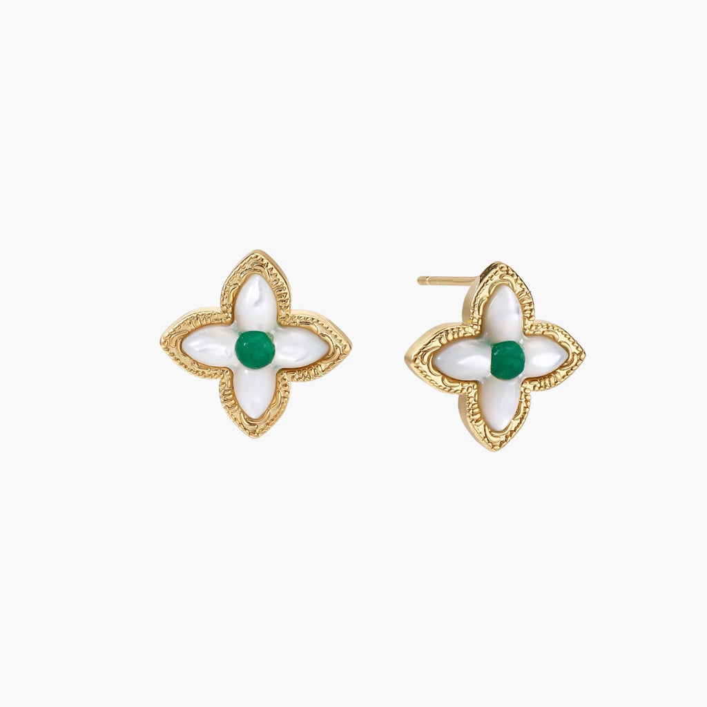 Gold quatrefoil stud earrings with white mother-of-pearl petals and emerald green gemstone center on beige background.