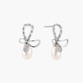 Silver bow-shaped stud earrings with baguette crystals and dangling white freshwater pearls on cream background