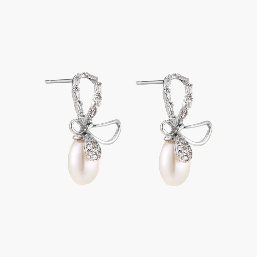 Silver bow-shaped stud earrings with baguette crystals and dangling white freshwater pearls on cream background