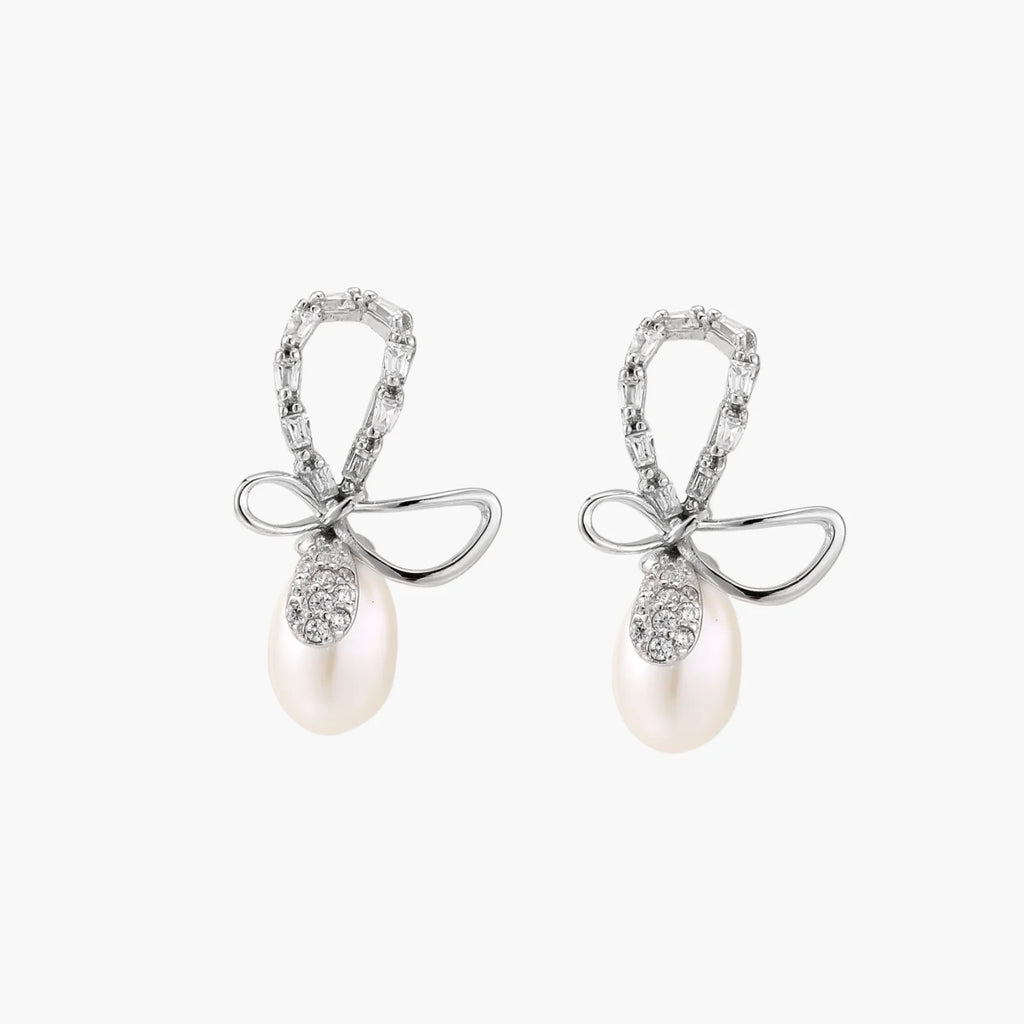 Silver bow-shaped stud earrings with baguette crystals and dangling white freshwater pearls on cream background