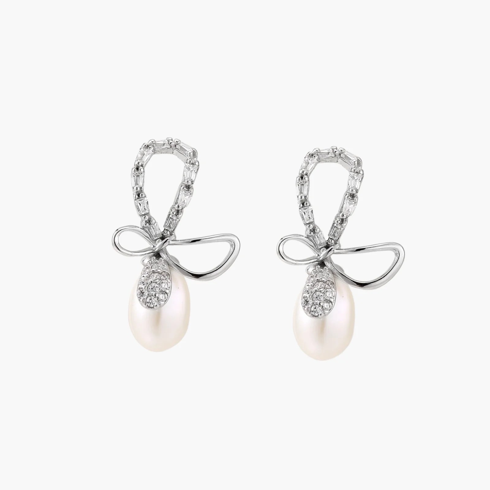 Silver bow-shaped stud earrings with baguette crystals and dangling white freshwater pearls on cream background
